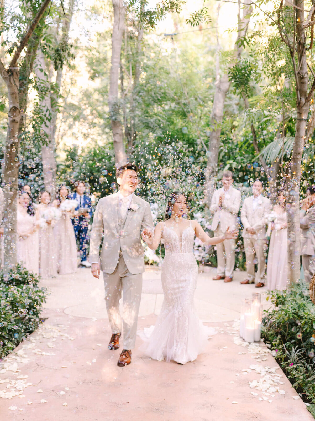 Bride and groom joyfully walk down a flower-petal-strewn aisle amidst bubbles. Wedding party claps in the background.