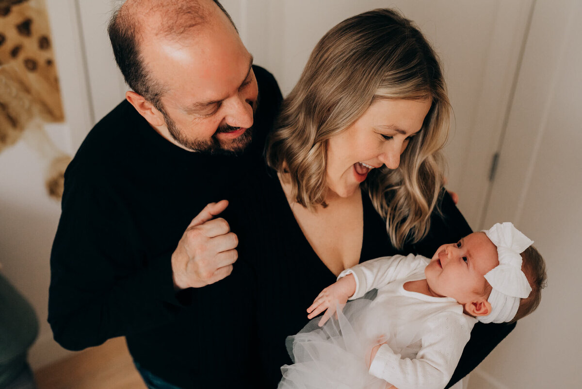 A mother and father hold their baby in their East Boston nursery, the baby smiles up at her parents during her photoshoot with Allison Wolf, Boston Newborn Photographer