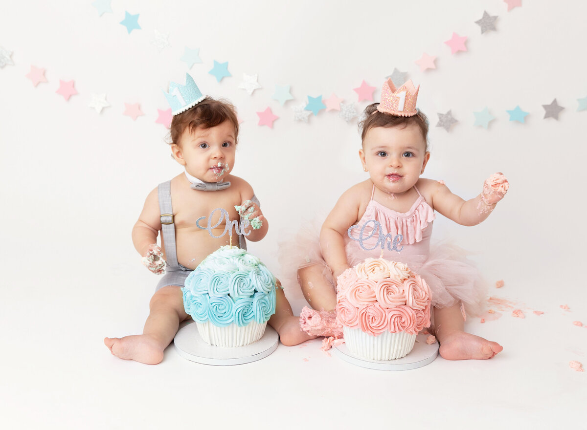 Twin boy and girl sitting for first birthday cake smash photos in Brooklyn, NY.  Baby boy has a blue cupcake cake and baby girl has a pink cupcake cake. Both babies have icing on their hands.