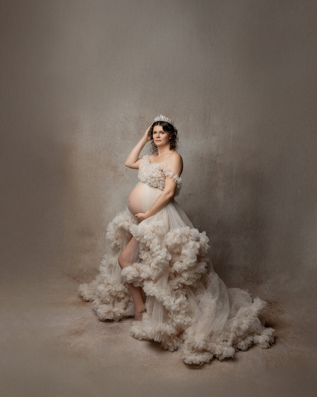 Graceful maternity portrait featuring a beautiful expectant mother in a voluminous, textured cream dress, gently embracing her growing belly against a muted, artistic background. Captured by Inland Empire maternity photographer Kaitlyn Dawn Photography.
