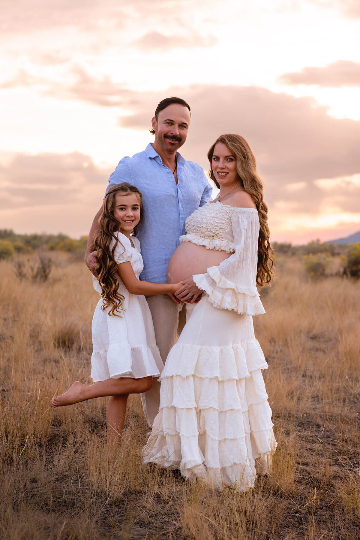 In Kamloops, Brittany Danielle captures a family maternity photo. Pregnant mom has long brown hair wearing a matching cream top and skirt with belly exposed. Husband stands behind and young daughter in fron with long brown hair. Young duaghter caresses moms belly and they all smile at the camera.