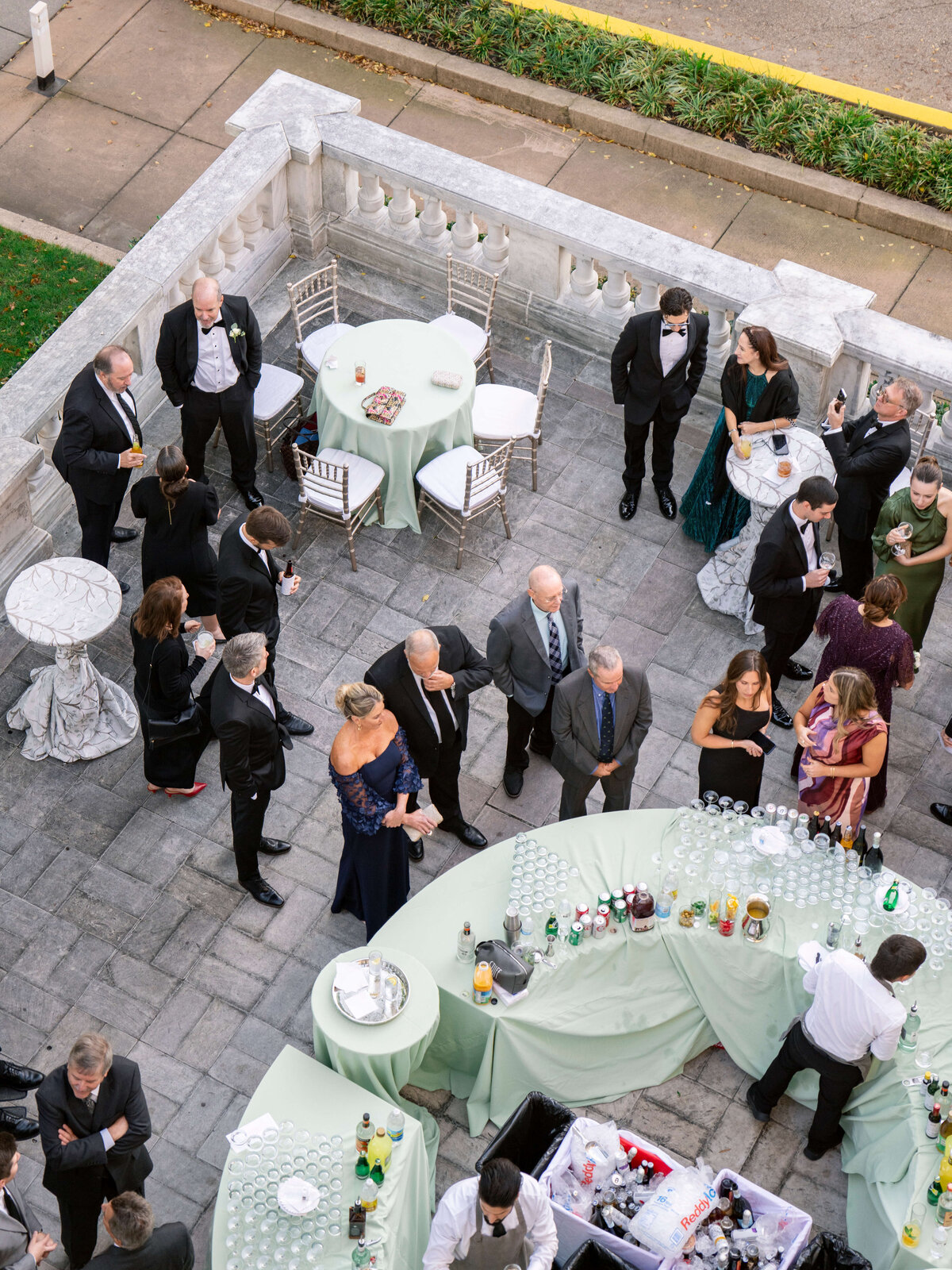 Aerial view of a formal gathering on a patio with people in evening attire. Guests socialize around tables with green tablecloths and a bar setup. White balustrades and decorative chairs are visible, creating an elegant atmosphere.