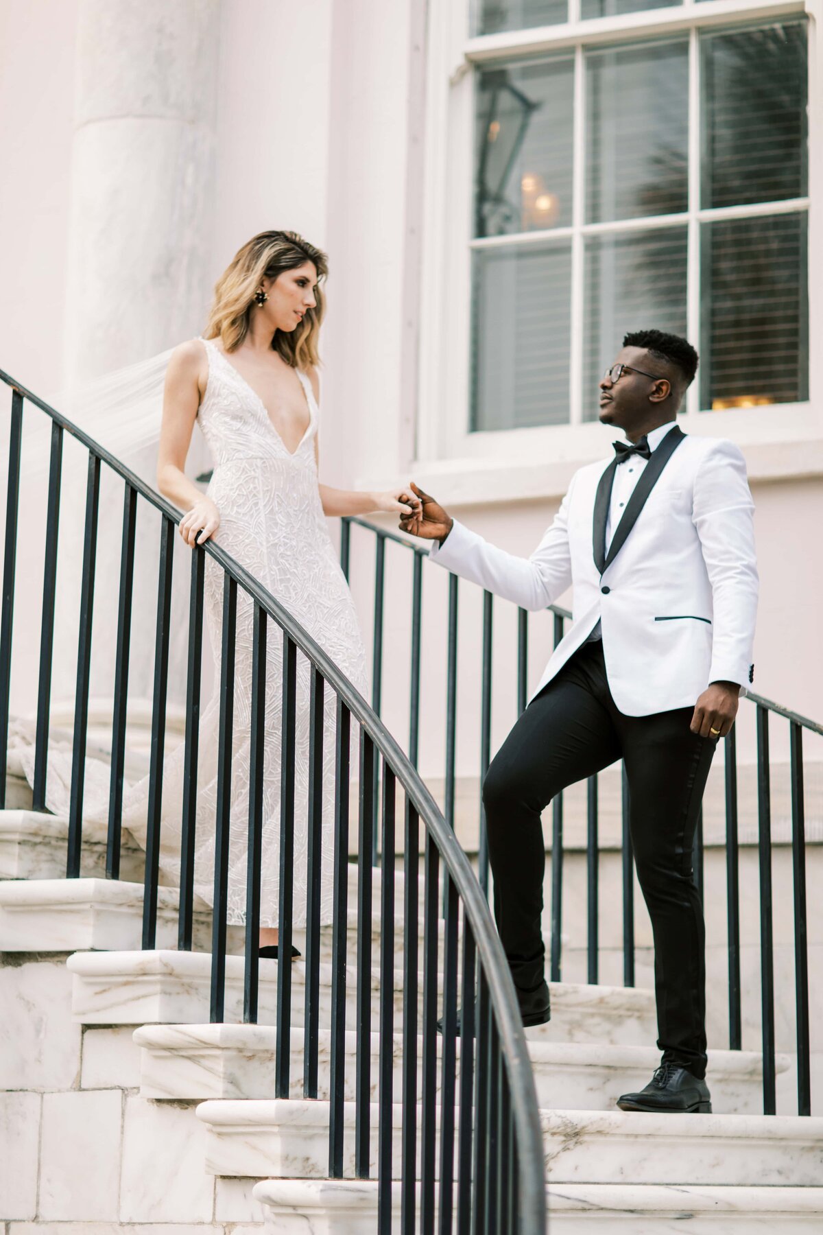 A beautiful white bride in a white wedding dress being escorted down the stairs by a handsome groom in a white and black tuxedo whilst looking at each other in Charleston