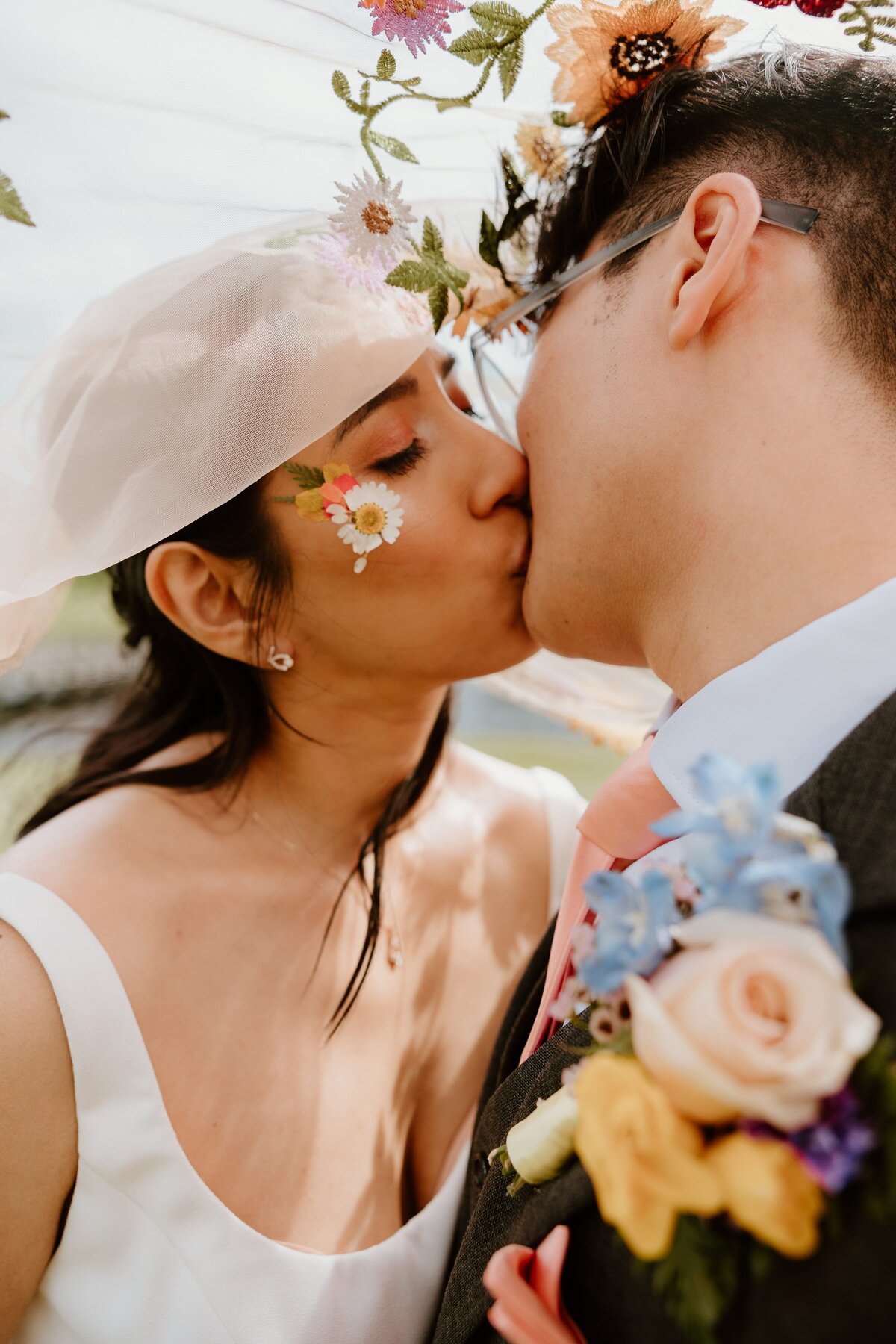 Close-up of bride and groom sharing a kiss beneath a floral-embroidered veil, with pressed flower details and colorful boutonniere at their romantic wedding at Hollow Brook Golf Club.