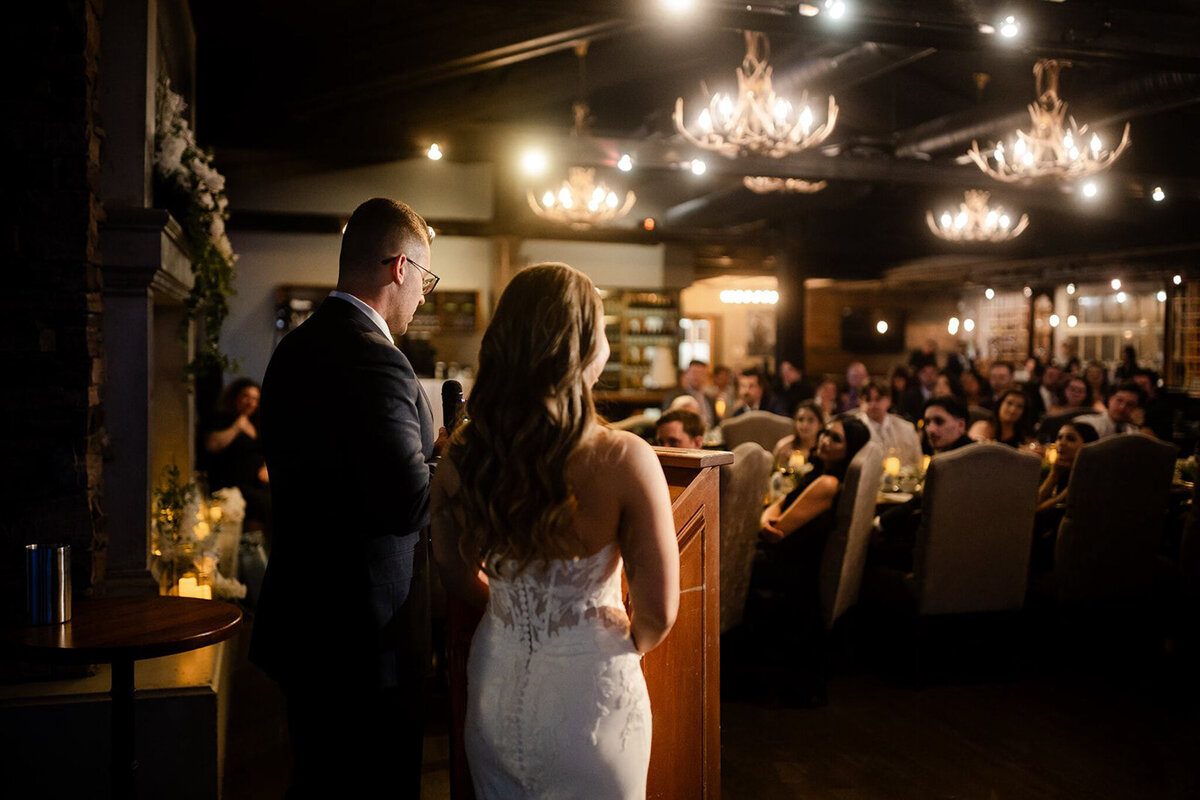 A warm and intimate wedding reception at The Lake House in Calgary, viewed from outside through large glass windows. Guests are seated at long, candlelit tables, engaged in conversation and enjoying the elegant atmosphere. The rustic charm of the venue is highlighted by stone walls, a grand fireplace adorned with greenery and candles, and antler chandeliers casting a soft glow. The dim lighting and reflections in the glass add depth to the scene, creating a cozy and romantic ambiance for the celebration.