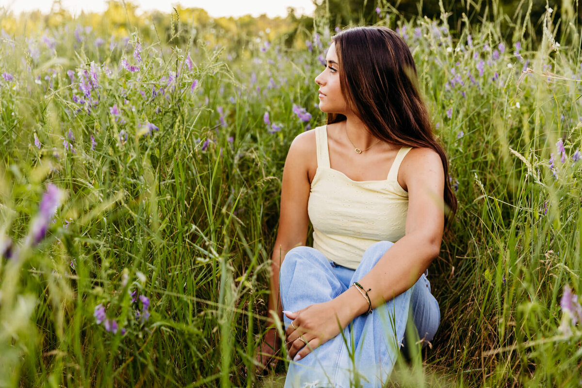 oshkosh high school senior girl sitting calmly in a field of purple flowers at sunset as she looks over her shoulder into the field captured by Ashley Kalbus Photography