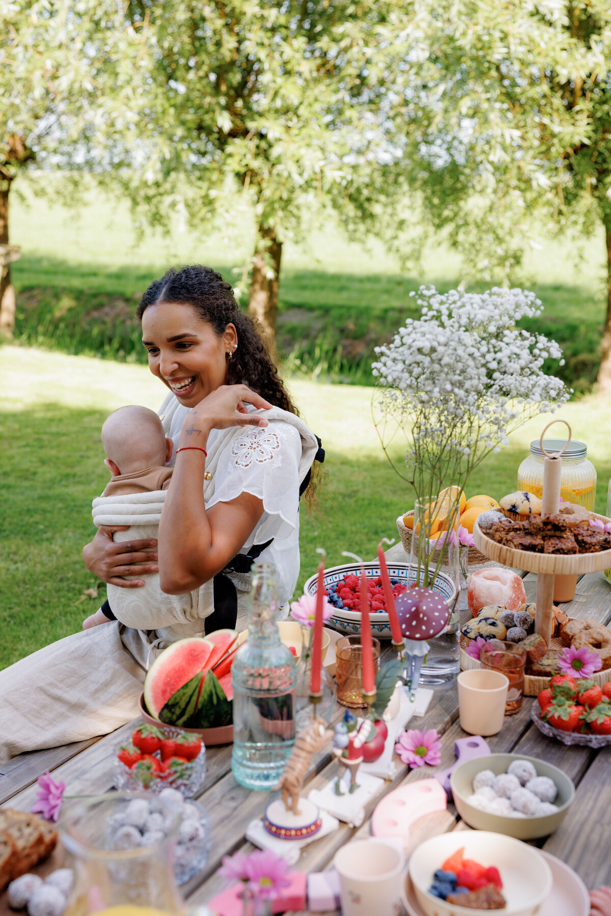 Vrouw met een baby geniet van een feestelijk gedekte picknicktafel in een groene omgeving bij Kinderdagverblijf Bij Ons Op De Boerderij.