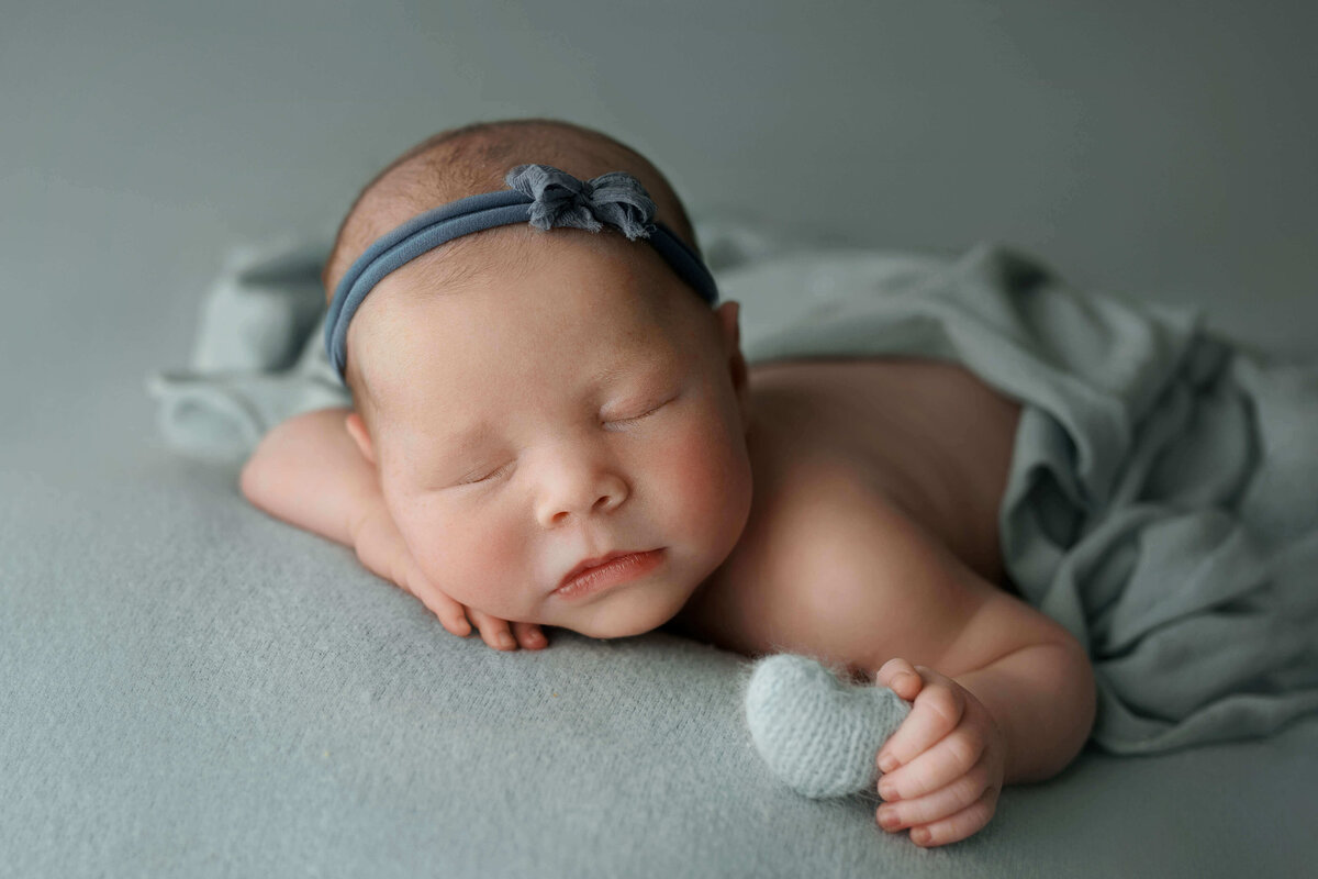 newborn baby girl laying on her tummy on a blue backdrop holding a knitted heart plushie at a photo shoot