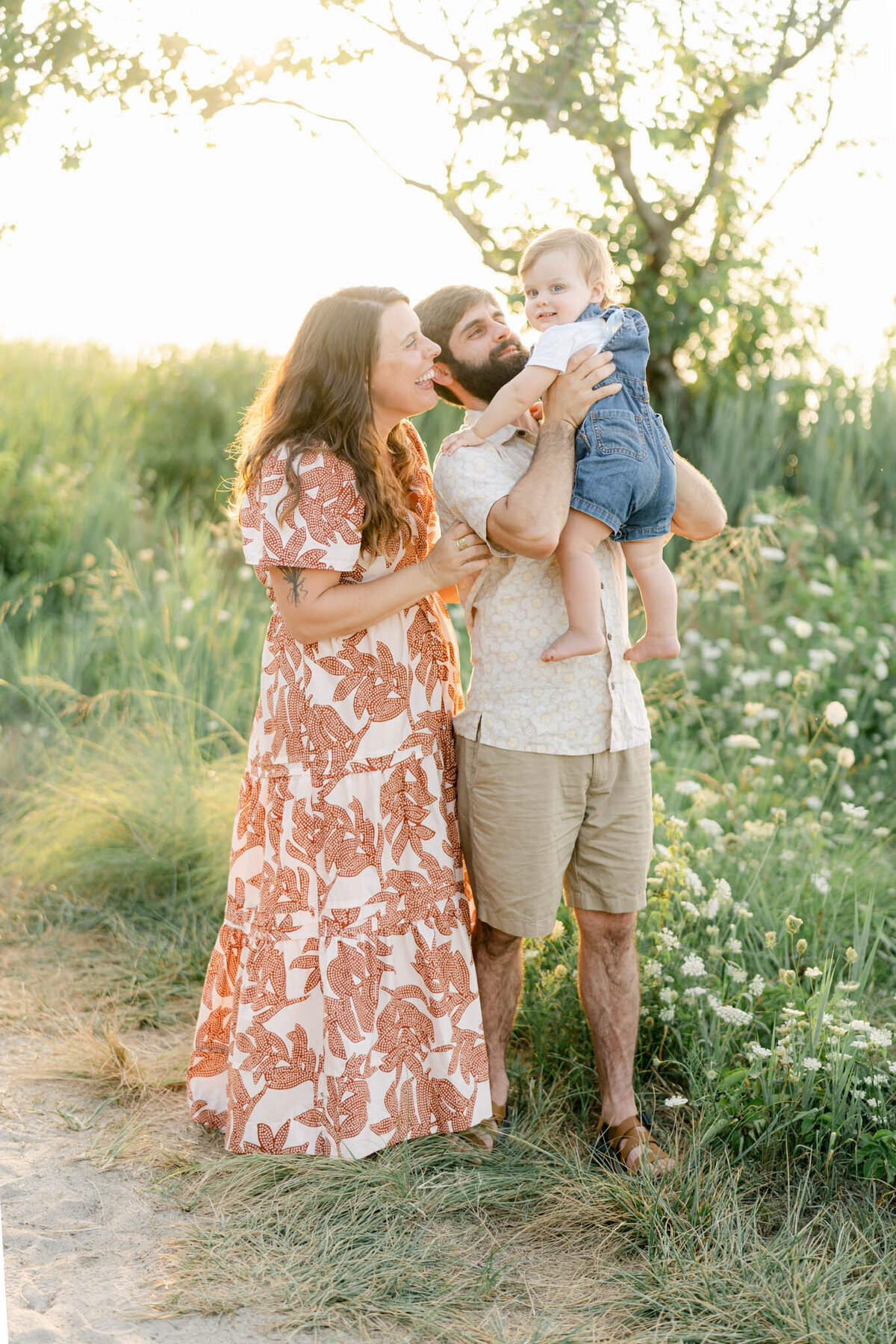 husband and wife hold baby boy in a field of wildflowers at sunset