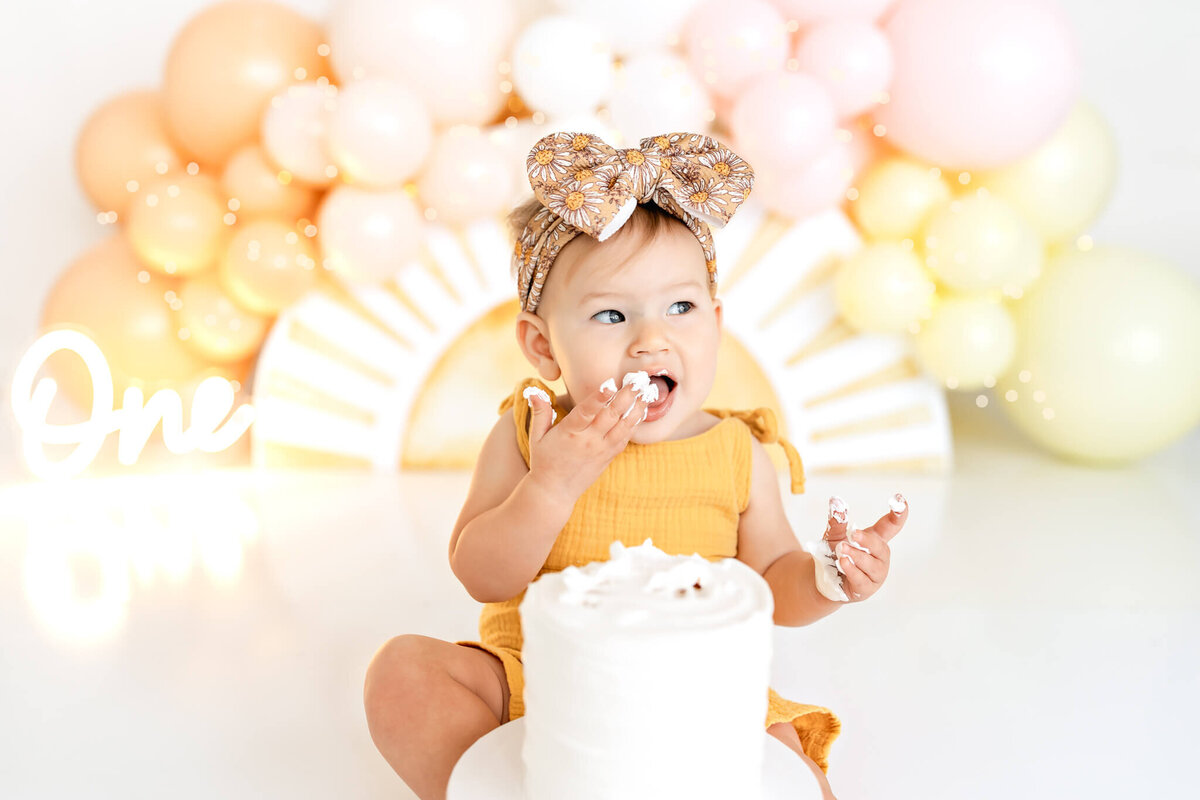 A baby wearing a yellow outfit and a bow sits on a glossy floor surrounded by pastel balloons. There's a partially eaten white cake in front of them, and "One" is displayed in white script in the background.