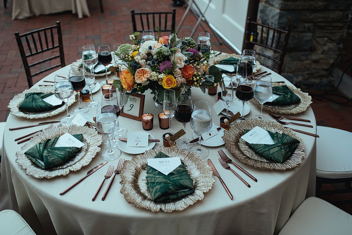 A round reception table with an ivory silk dupioni table cloth with gold glass chargers and menu cards wrapped in forest green napkins with gold flatware and a large orange, pink, white purple and green floral centerpiece and brown chiavari chairs