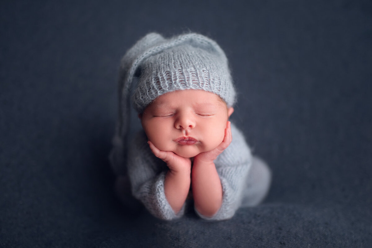 Blue-themed portrait of newborn baby boy wearing blue stocking cap and matching onesie. He is asleep on his tummy  with his chin in his hands.