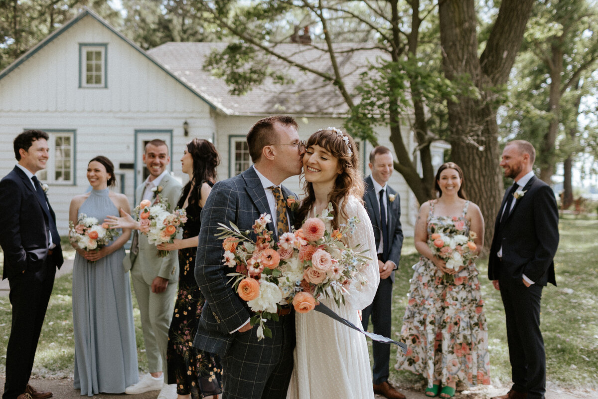A bride and groom stand smiling with flowers, surrounded by a happy bridal party outdoors at the Queen City Yacht Club, near a white house and trees, all beautifully captured by Jennifer van Son, a talented wedding photographer in Toronto.