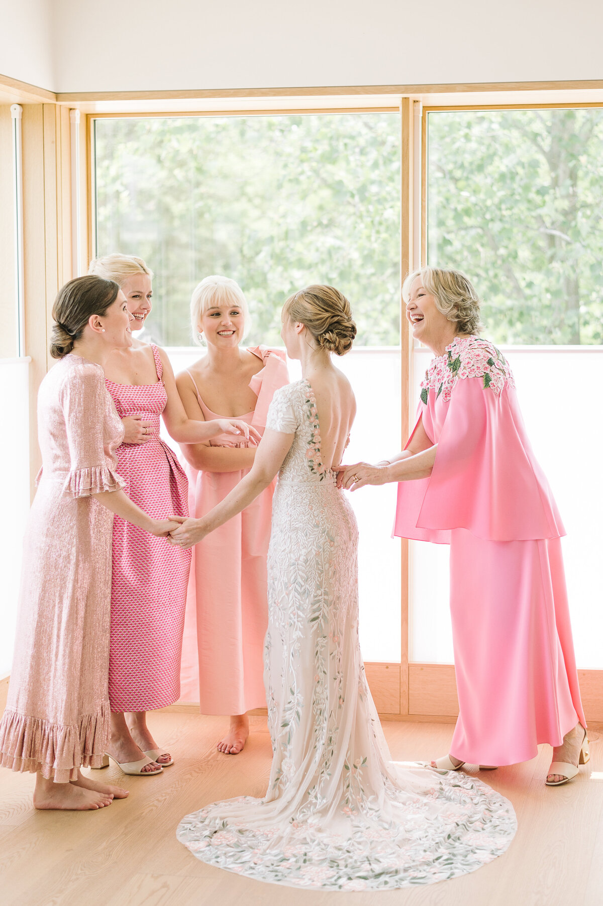 Sisters and mom of the bride all dressed in pink help the bride to put on her wedding dress
