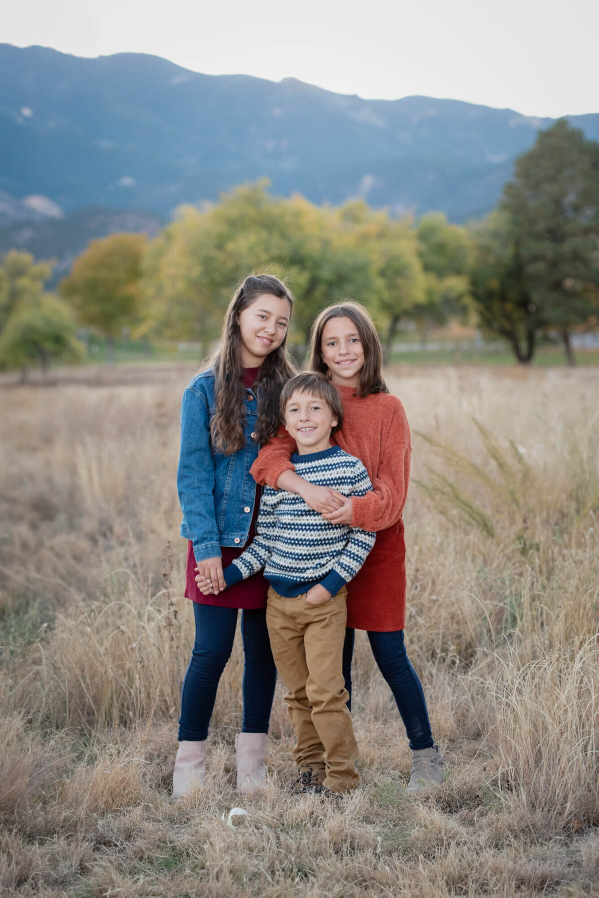 3 young siblings hug and hold hands while smiling in a park trail for a Colorado Springs family photographer