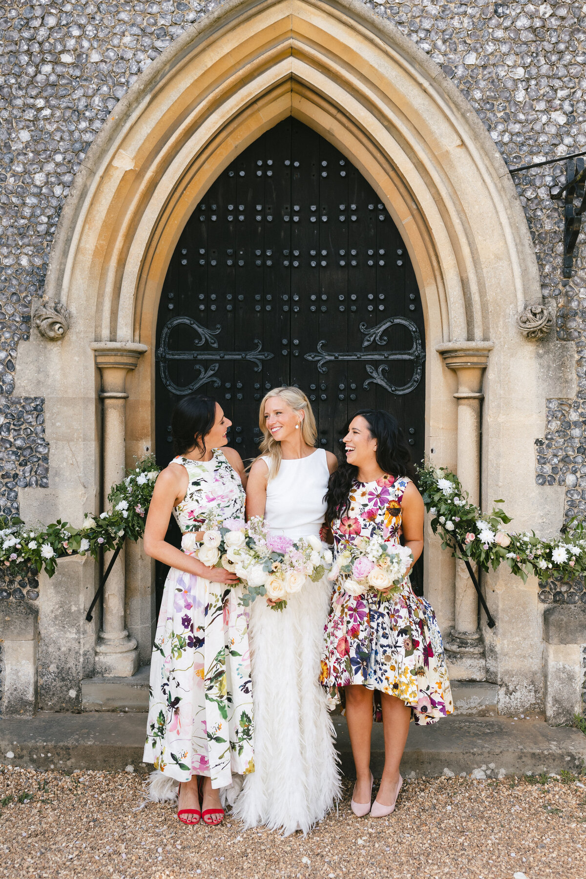 A Bride with her bridesmaids stand smiling in front of an arched doorway decorated with flowers. The woman in the middle wears a white dress, while the others wear floral dresses and hold bouquets. The setting has a stone facade and black double doors.