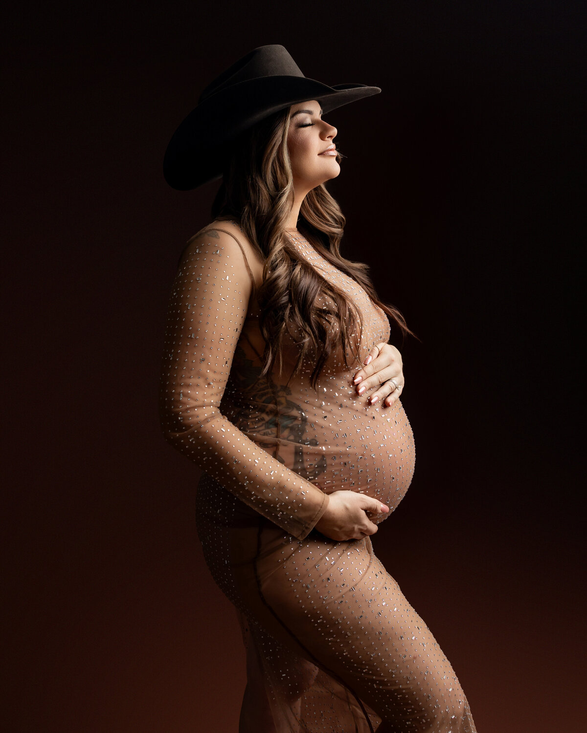 A glowing expectant mother wearing a cowboy hat and a sheer brown gown, set against a dark, moody backdrop. Captured by Inland Empire maternity photographer Kaitlyn Dawn Photography.