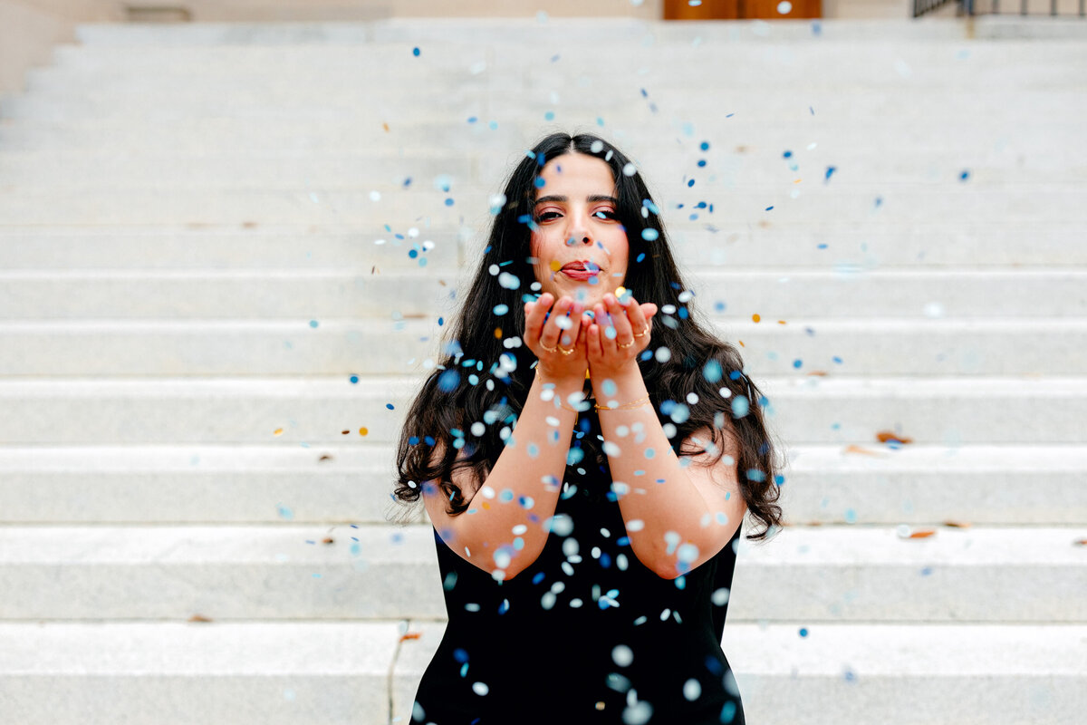 UNC graduate in black dress celebrating by blowing confetti at Wilson Library.