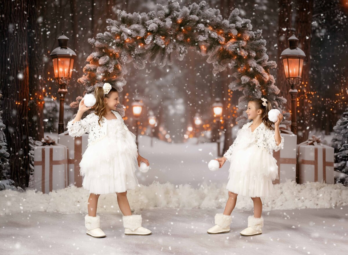 Two girls in white dresses and boots throwing snowballs at each other. Holiday mini sessions photographed at Atlanta photo studio. 