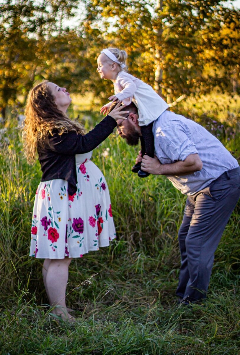 a pregnant woman blows kisses to her daughter who is on her dad's shoulders