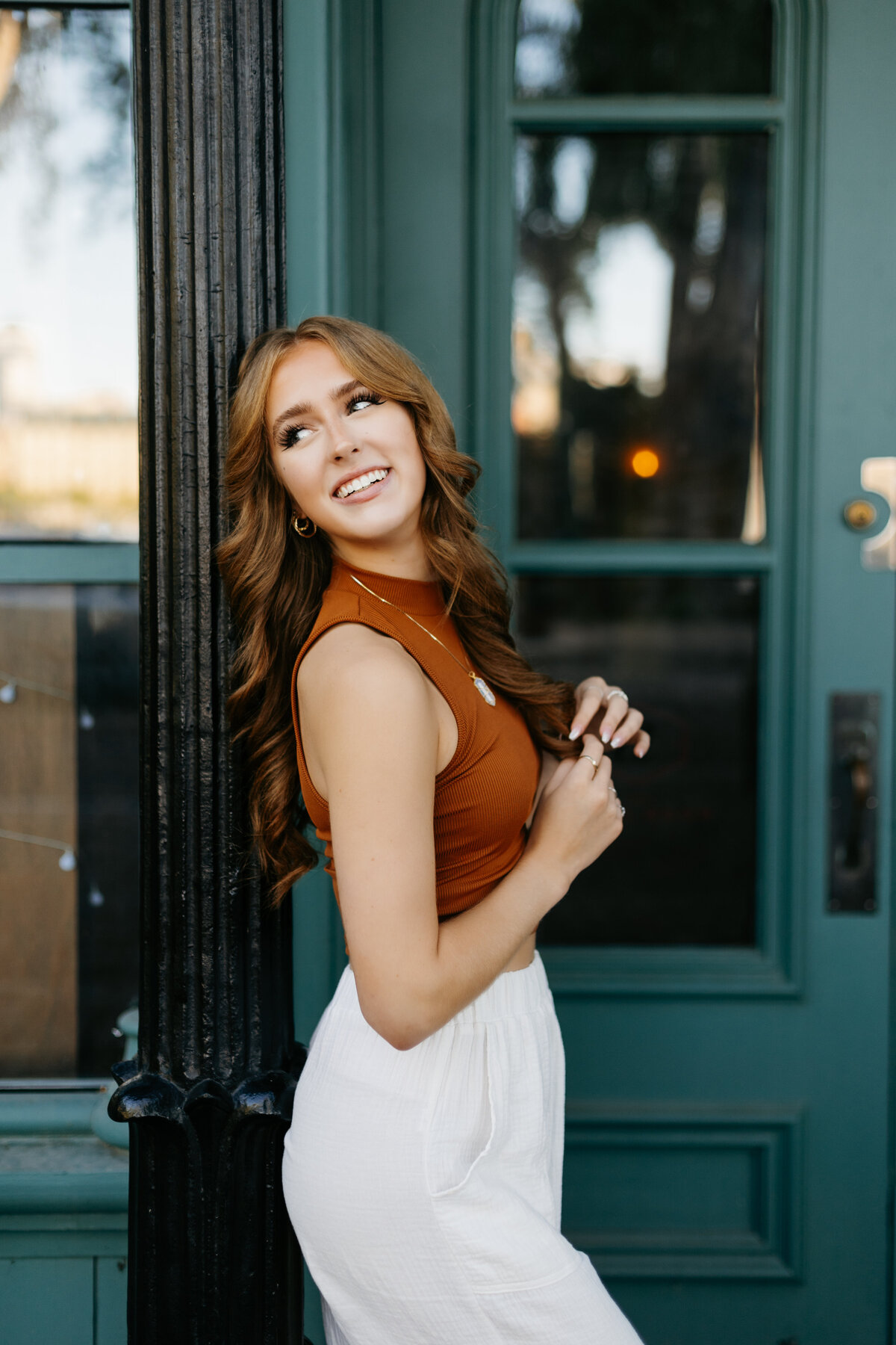 Senior girl in a brown top and white pants leaning against a pole in front of a green door