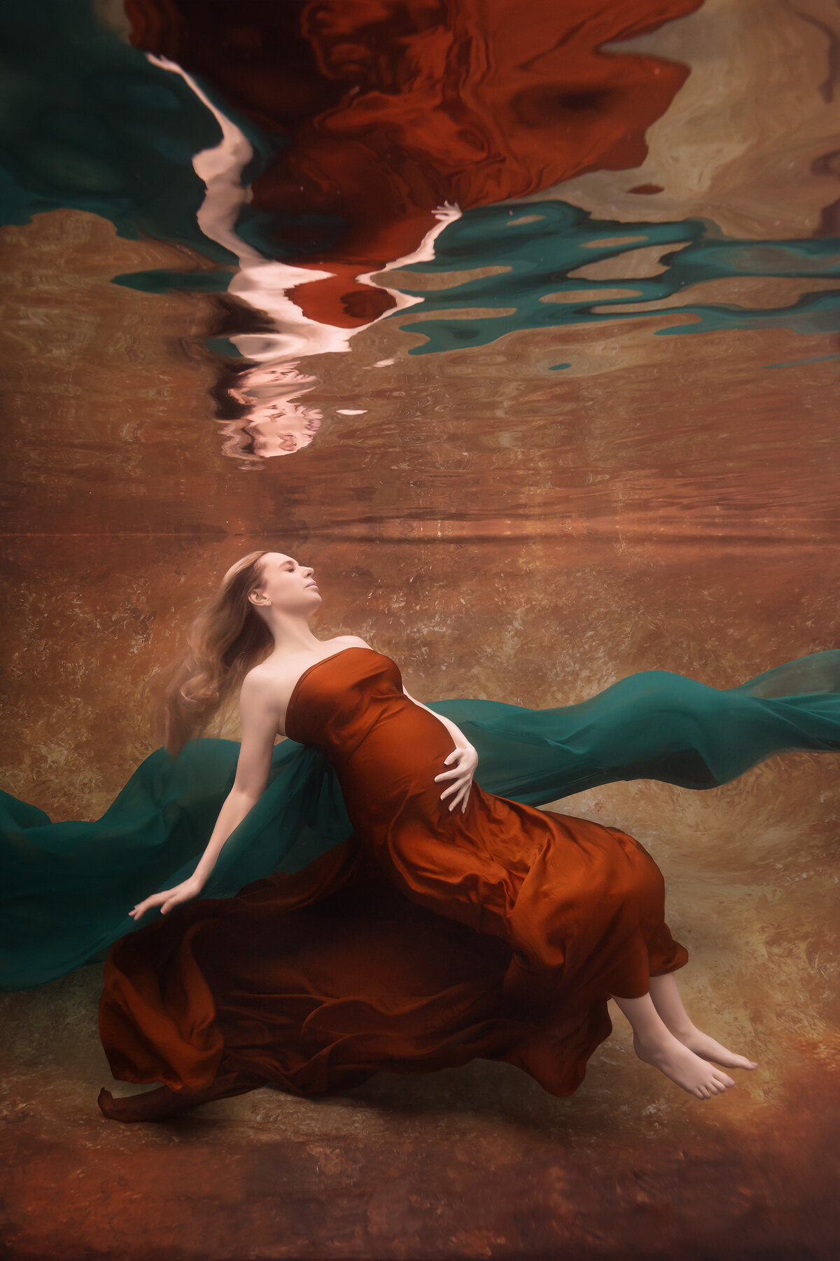 An ethereal underwater portrait captured by Lindsay Walden Photography, featuring a woman gracefully reclining in a rust-colored gown against a golden backdrop. Flowing emerald fabric swirls around her, enhancing the luxurious feel of this fine art underwater maternity photoshoot.
