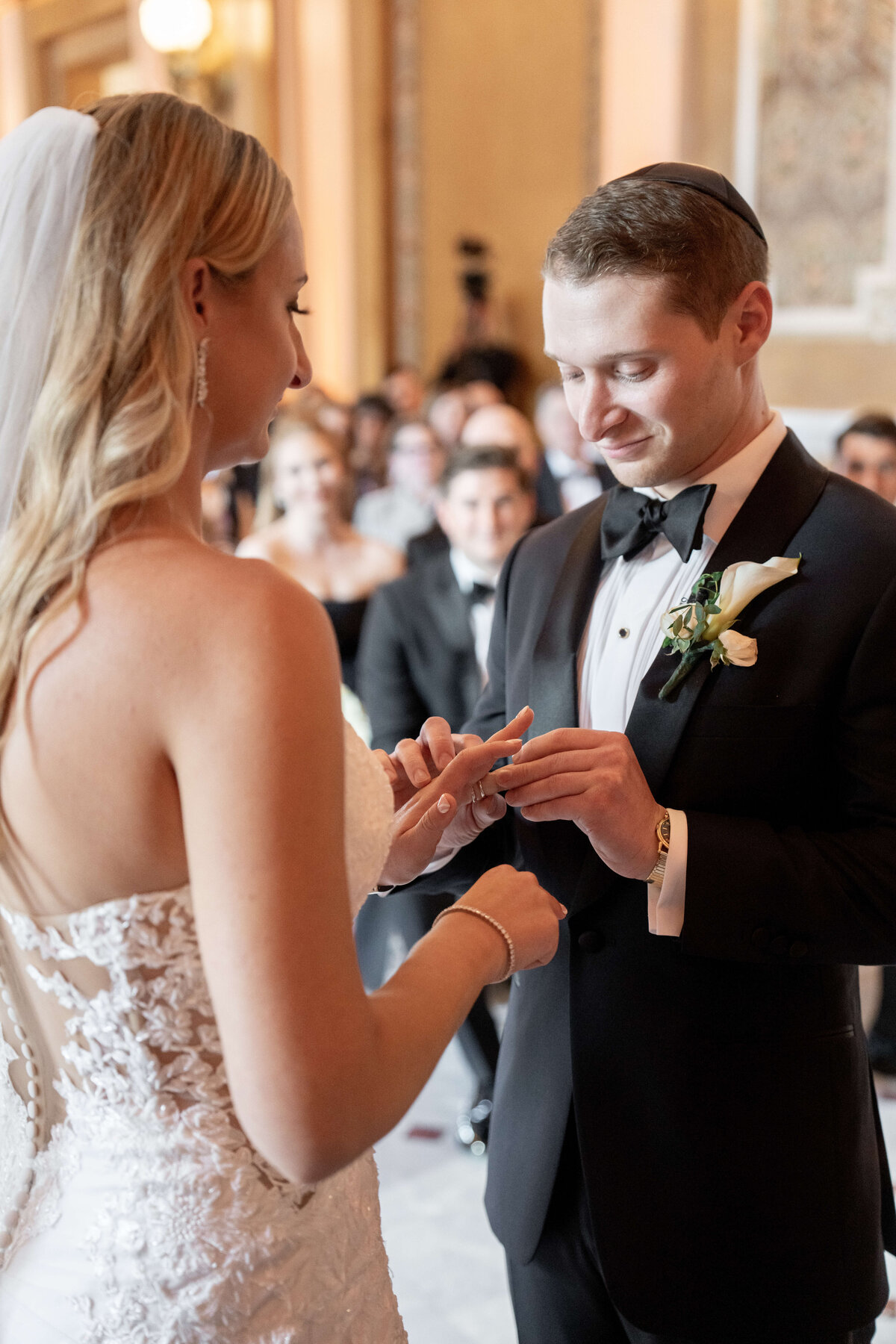 A bride and groom exchange rings during a wedding ceremony. The bride is in a white lace gown and veil, while the groom wears a black tuxedo with a white boutonniere and a kippah. Guests are seated in the background.