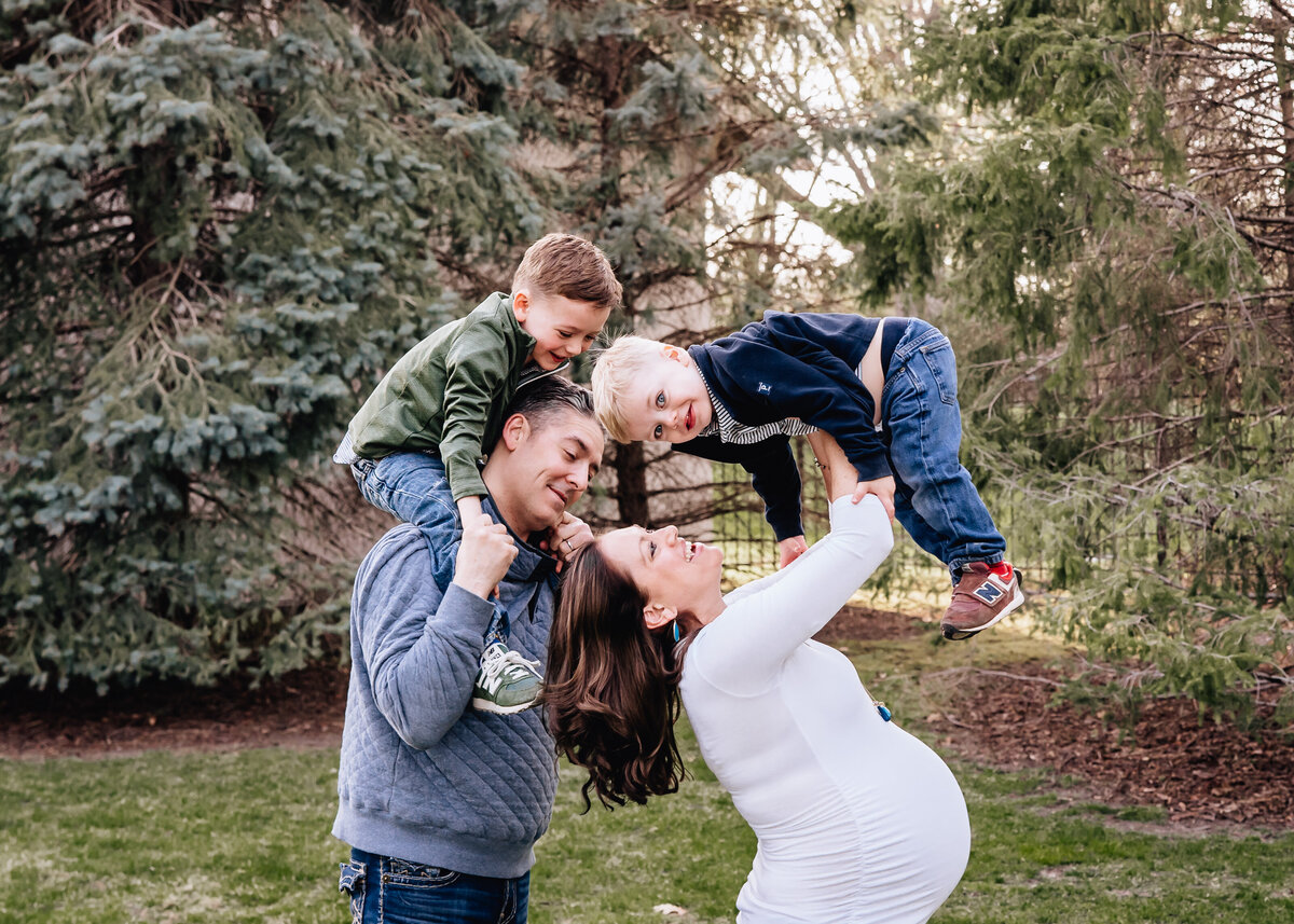 A family of four enjoys a playful moment outdoors. The mother, pregnant and smiling, lifts one child in the air while the father carries the other child on his shoulders.