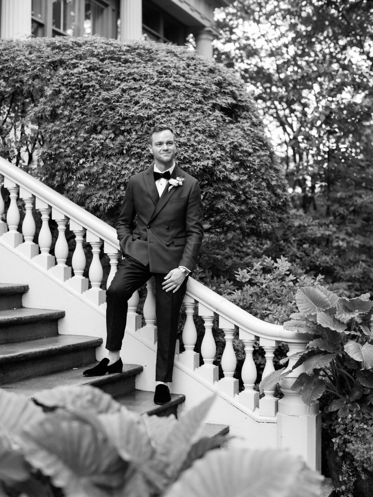A dashing groom poses gracefully on a grand staircase adorned with lush greenery, embodying elegance and charm, perfect for capturing the essence of wedding attire and style.
