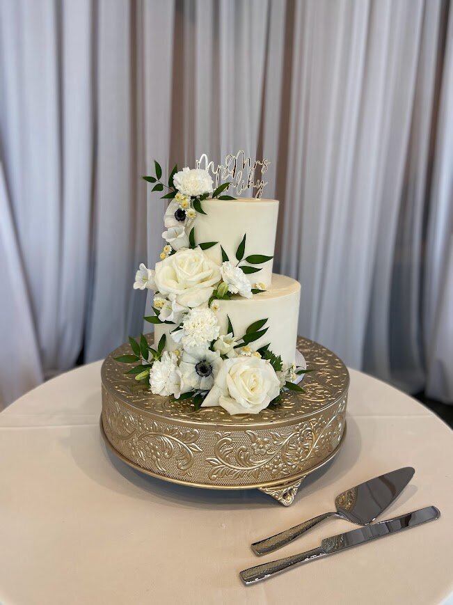 A white two tier cake sits on a gold cake stand. The frosting on the cake is smooth, and there's lots of white flowers and greenery cascading down the left side and across the front.