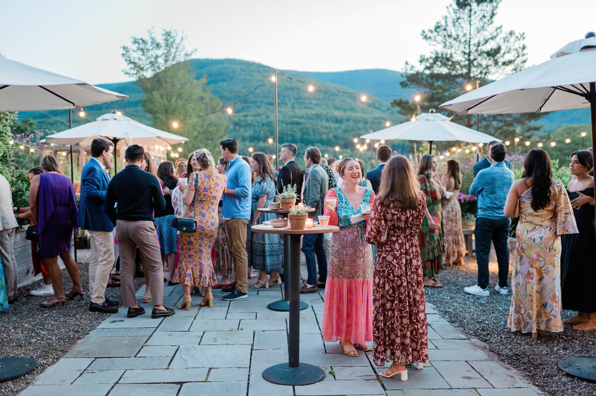 A crowd of well dressed guests chats and laughs under string lights on a patio, with mountain views and decorative umbrellas.