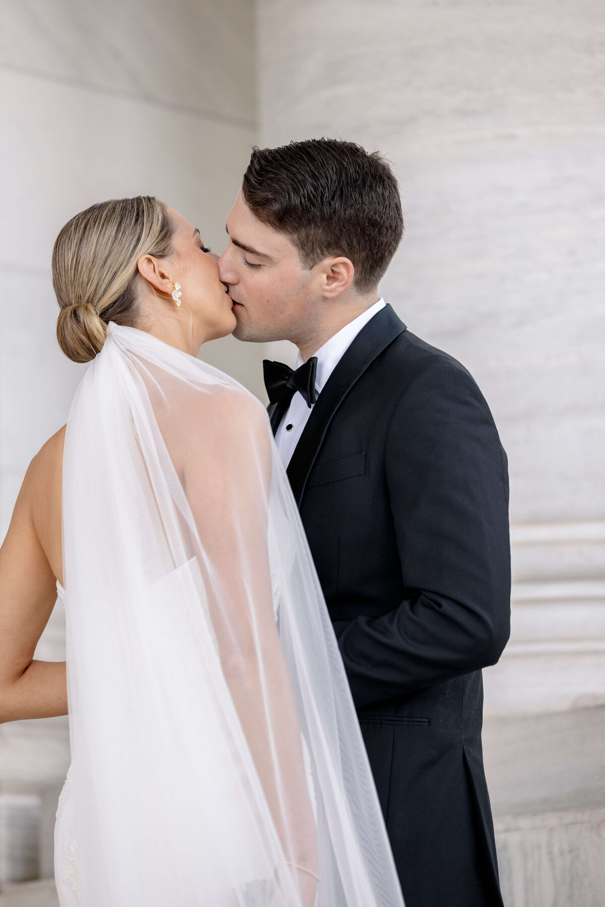 A bride and groom share a kiss. The bride wears a white gown with a sheer veil, and her hair is styled in an elegant bun. The groom is in a black tuxedo. They are standing in front of a stone architectural backdrop.
