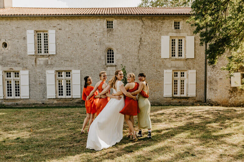 Mariée de dos entourée de deux demoiselles d'honneur de chaque côté, bras dessus, bras dessous, regardent en arrière. Bâtiment ancien en arrière plan. Photo immortalisée par Laura, photographe professionnelle en vendée.