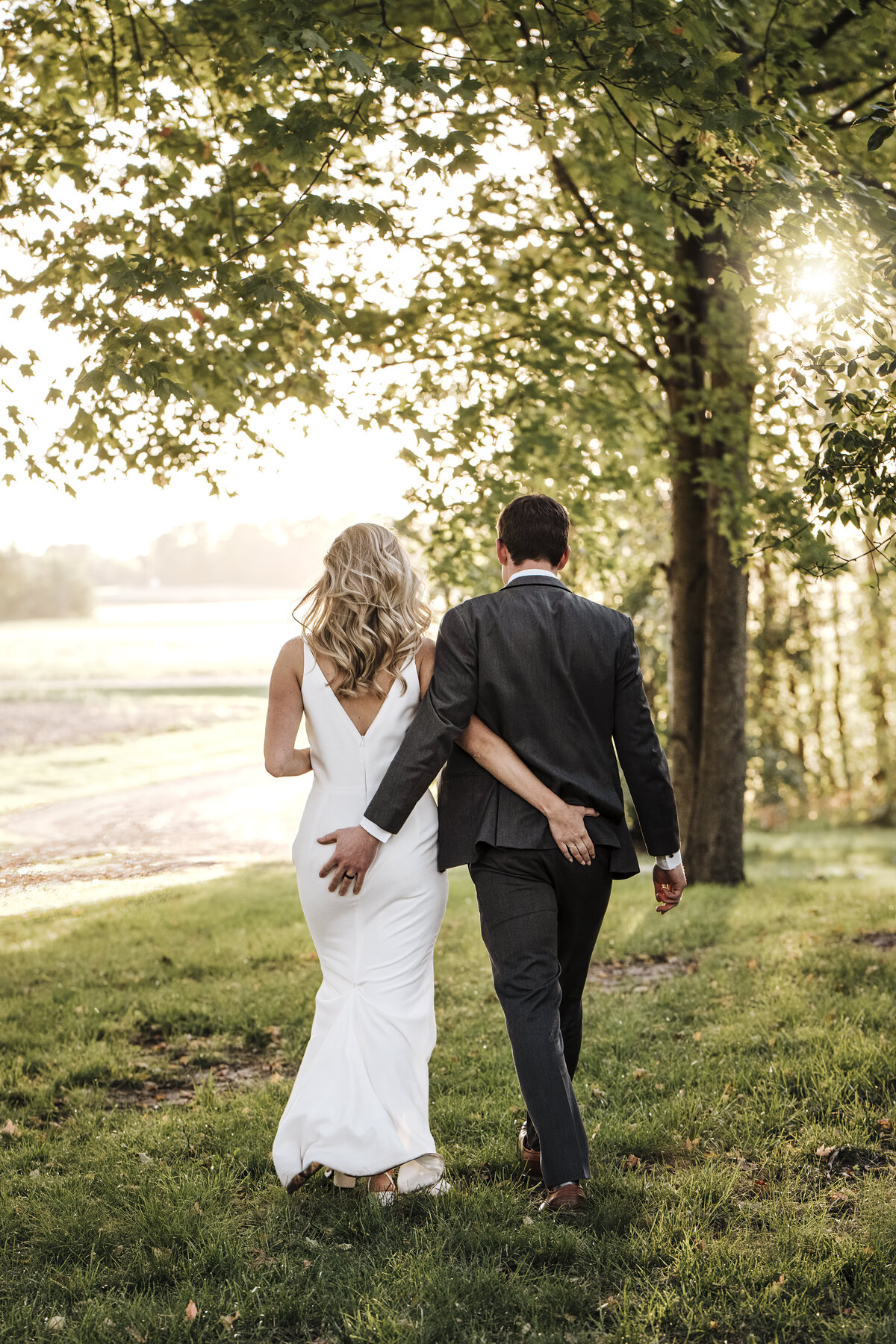 A couple in wedding attire walking hand-in-hand through a tree-lined path with hands on each others butt, with the sun casting a warm glow through the leaves taken by jen Jarmuzek photography a Minneapolis wedding photographer
