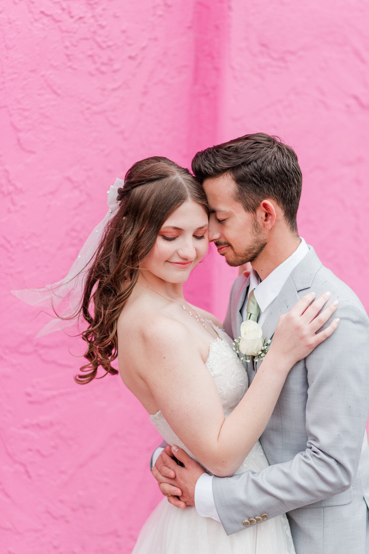 A bride and groom embrace with the groom's arms around his bride's waist and her hand at his shoulder while she gazes downward in front of a bright pink wall in downtown Denver