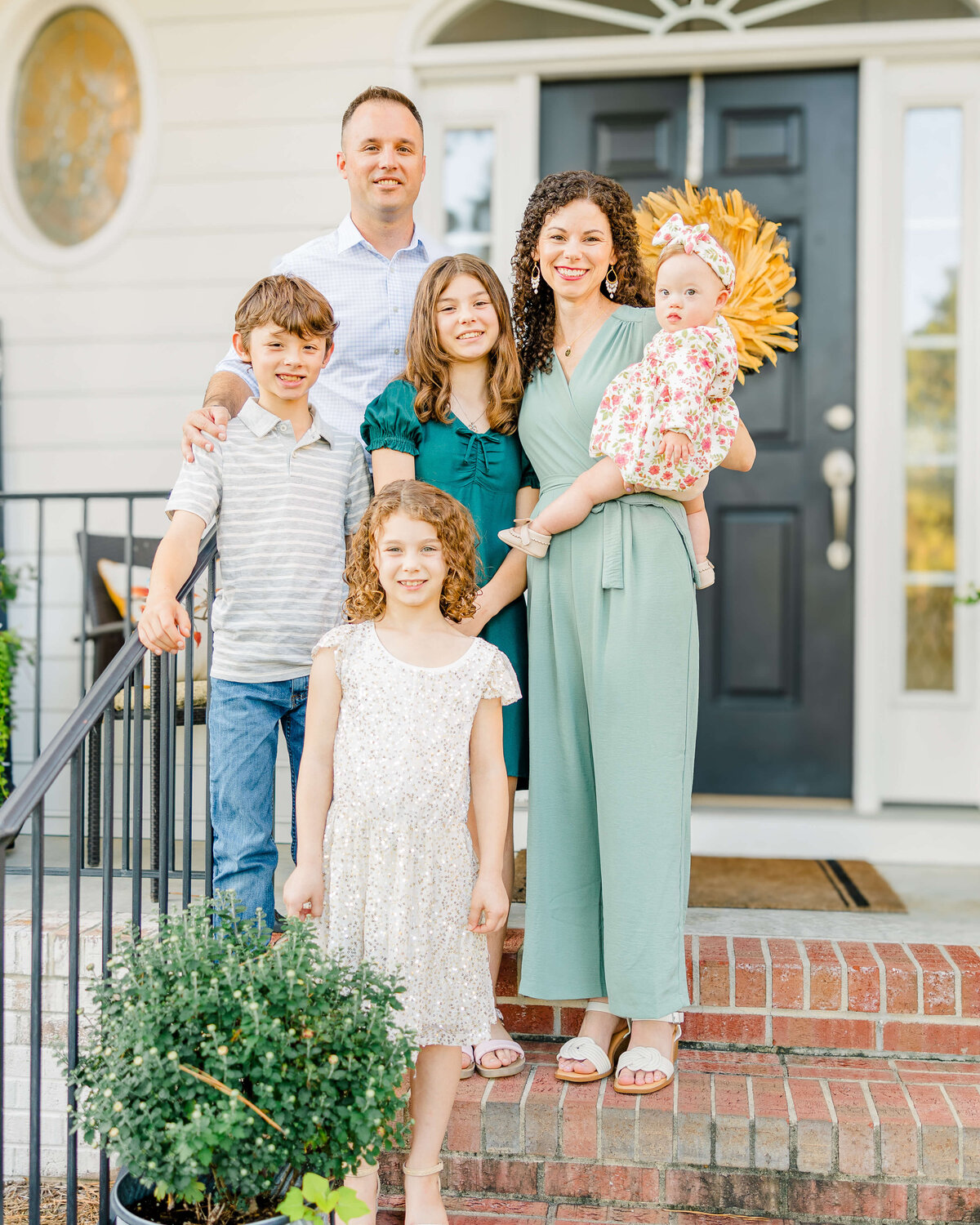 A family of six poses warmly on the porch, captured perfectly by a Fayetteville NC portrait photographer. The mother cradles a baby, surrounded by two girls and a boy, while the father beams behind them. A black door with a wreath and a potted plant adorns their charming home.