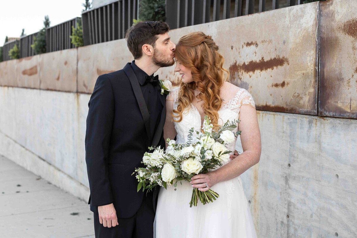 Groom holding bride and kissing her on the forehead while bride holds wedding bouquet