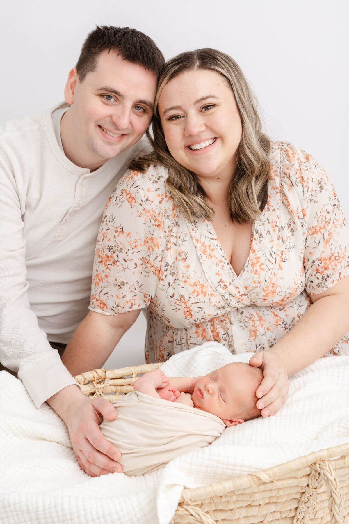 New Mom and New Dad are  sitting behind a Moses Basket with their newborn baby who is wrapped in a beige swaddle. Mom has her hand on baby's head and dad has his hand on baby's bottom. Mom and dad are sitting close together with their foreheads touching and they are both smiling proudly at the camera.