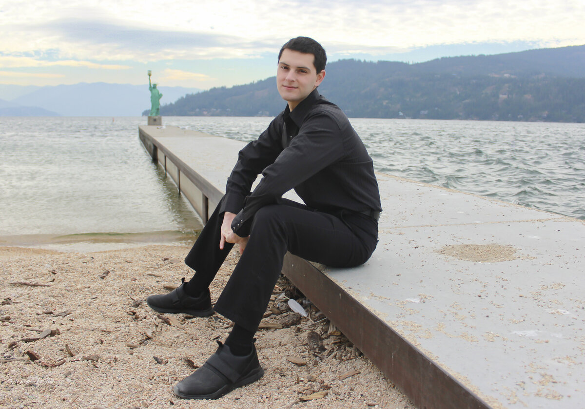 photo of a Man sitting on a pier  near lake with a statue of liberty in background