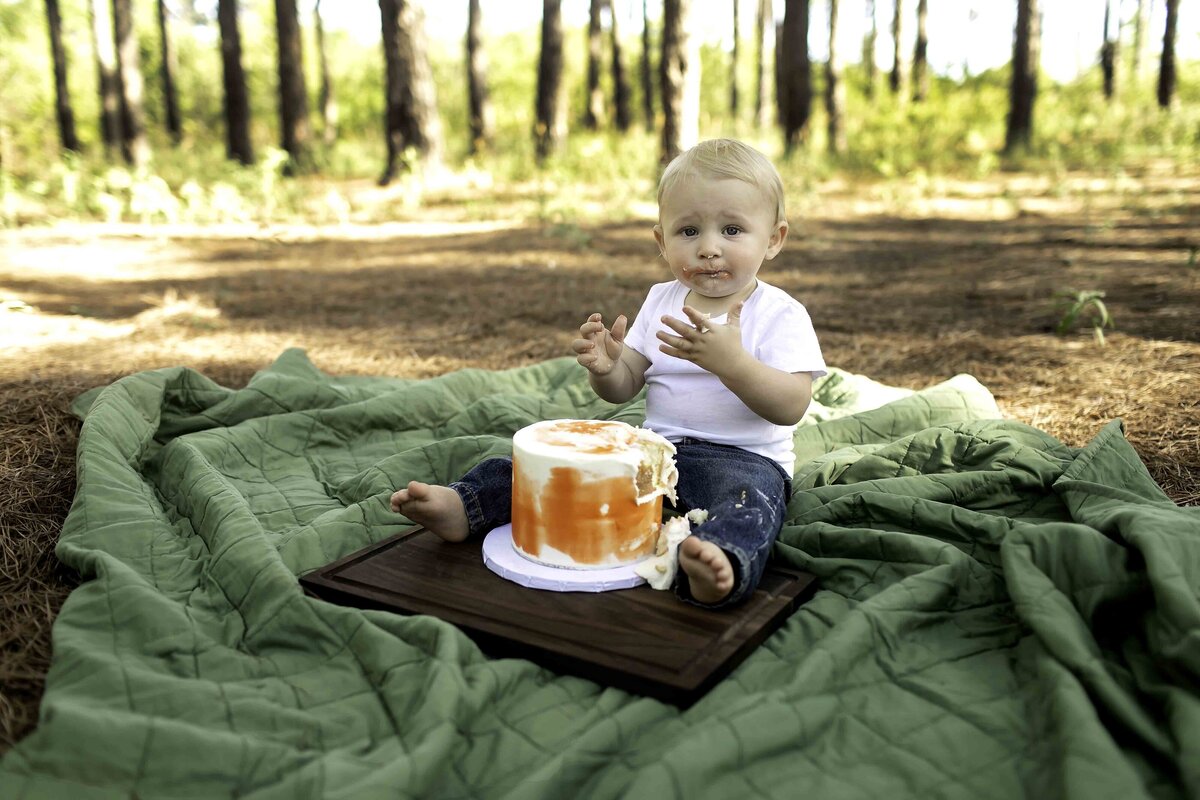Birthday boy in suspenders and overalls, covered in cake frosting, playfully smashing his birthday cake under the pine trees