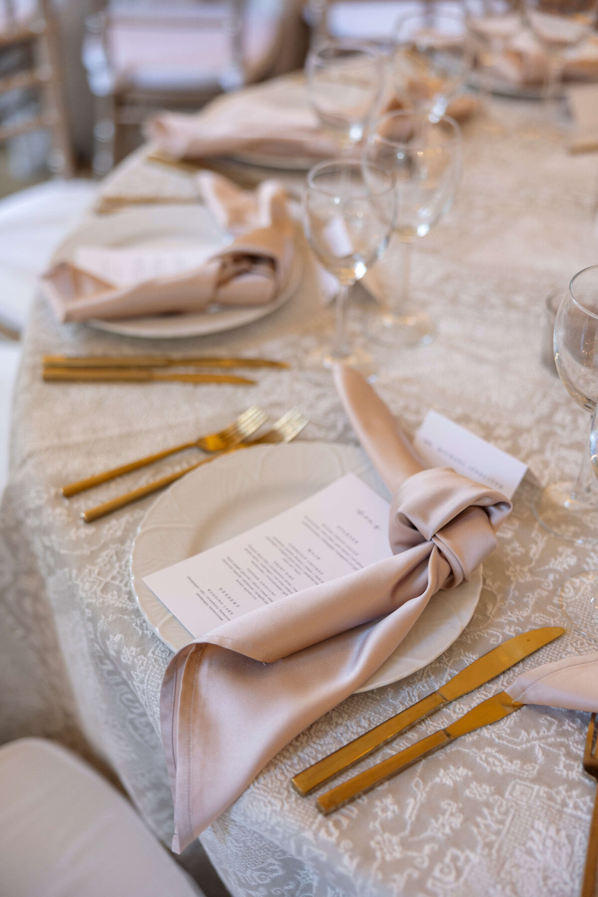 Elegant table setting with beige napkins gracefully folded over white plates. Gold cutlery is neatly arranged alongside a printed menu card. Glasses are placed above the plate on a light beige tablecloth featuring an intricate pattern.