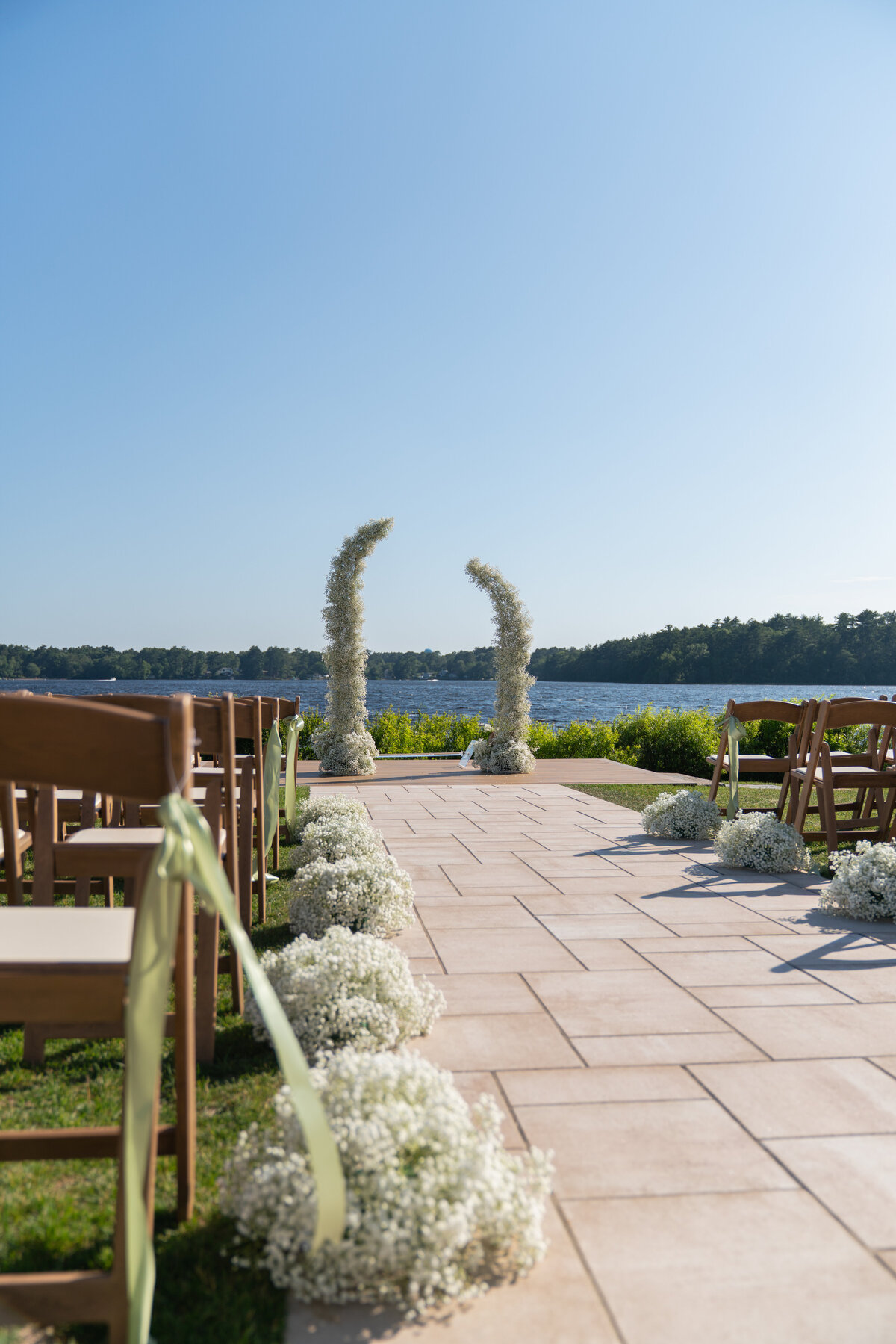 The Lakehouse Inn MA  | Kelsey Sheehan Photography Timeless Rhode Island Weddings | Outdoor wedding setup by a serene lake, with wooden chairs and floral arrangements along a tile aisle. Two tall flower arches frame the blue sky backdrop.