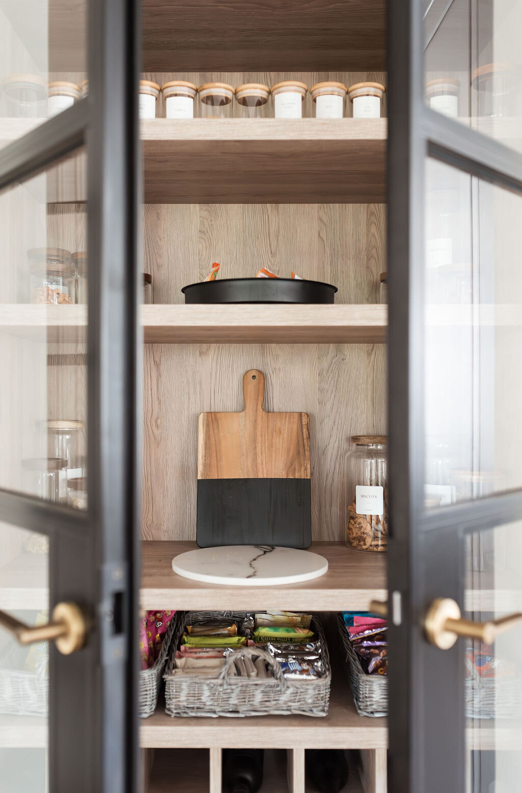 Built-in pantry cabinet with glass doors with black frames. Inside the open doors, are organized snacks and cutting boards.