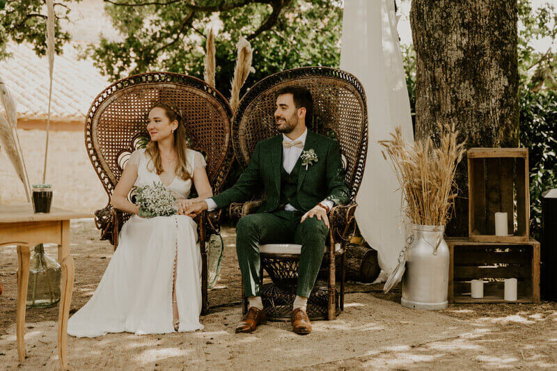 Mariés assis dans des fauteuils vintage et se tenant la main lors de la cérémonie laïque en extérieur. Photo par Laura Termeau, photographe professionnelle en Vendée.