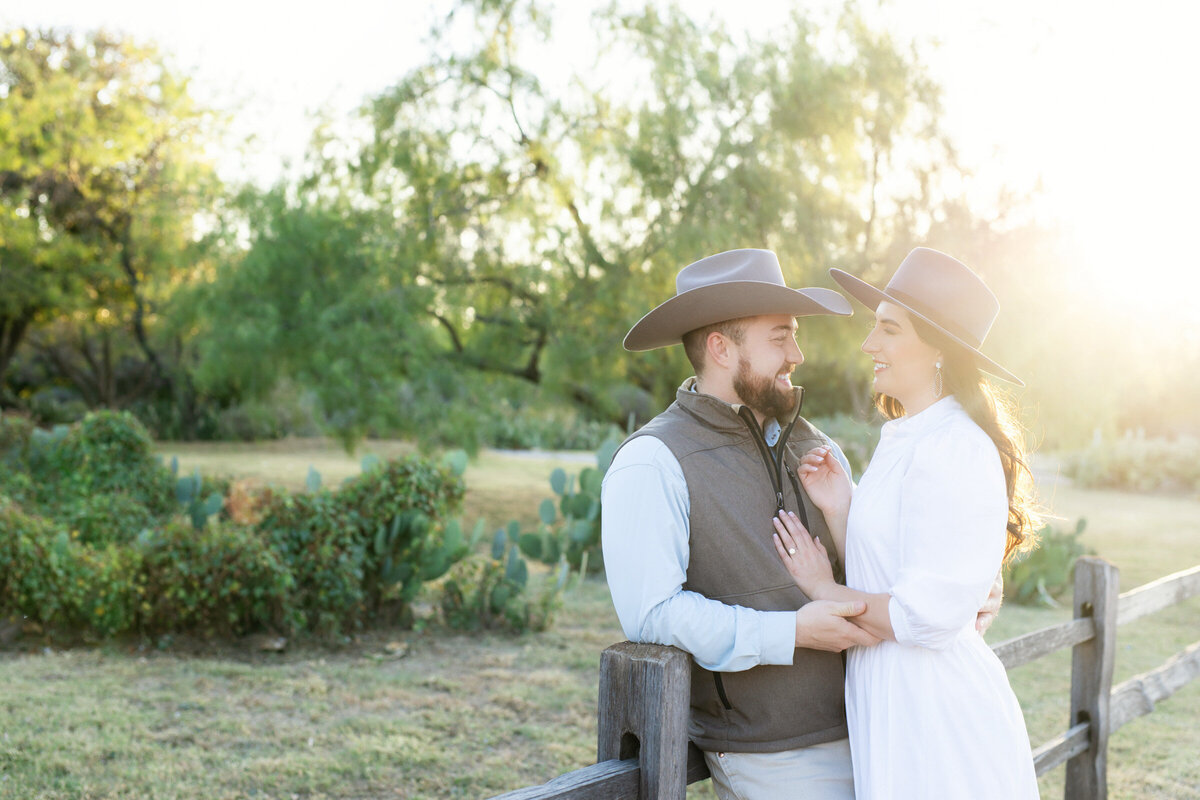 Engaged couple at the Drover Hotel in Fort Worth during sunset, he leaning on a fence while she stands close, both wearing cowboy hats