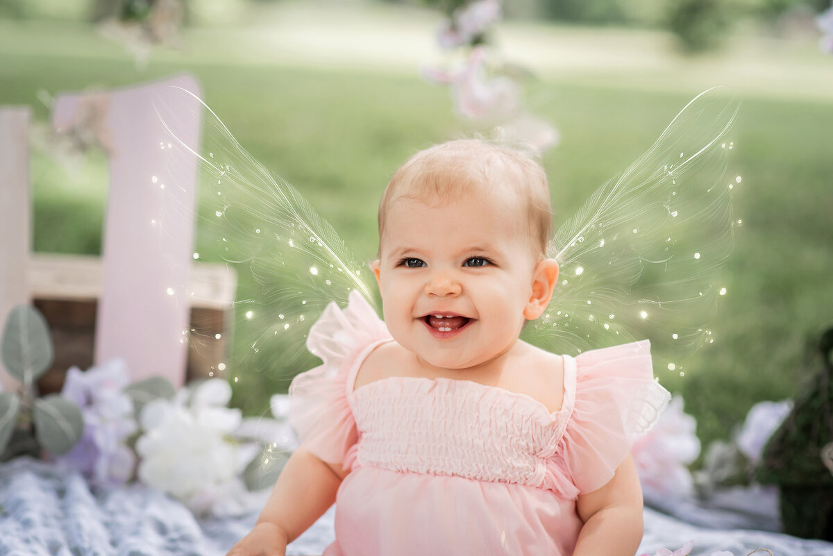 One year old girl with fairy wings laughing  white sitting on the grass in front of a pink wooden one