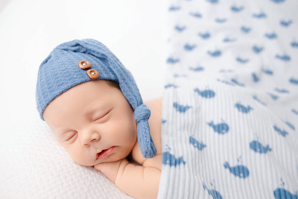 Newborn baby boy wearing a blue sleepy bonnet and covered over his back with a blanket at Heather Nicole Photography.