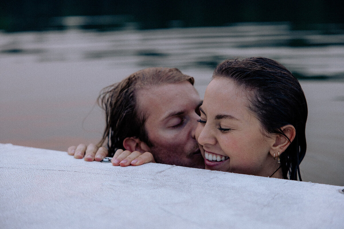 Boy kisses girl on the cheek as both are seen holding onto a dock in the water. 