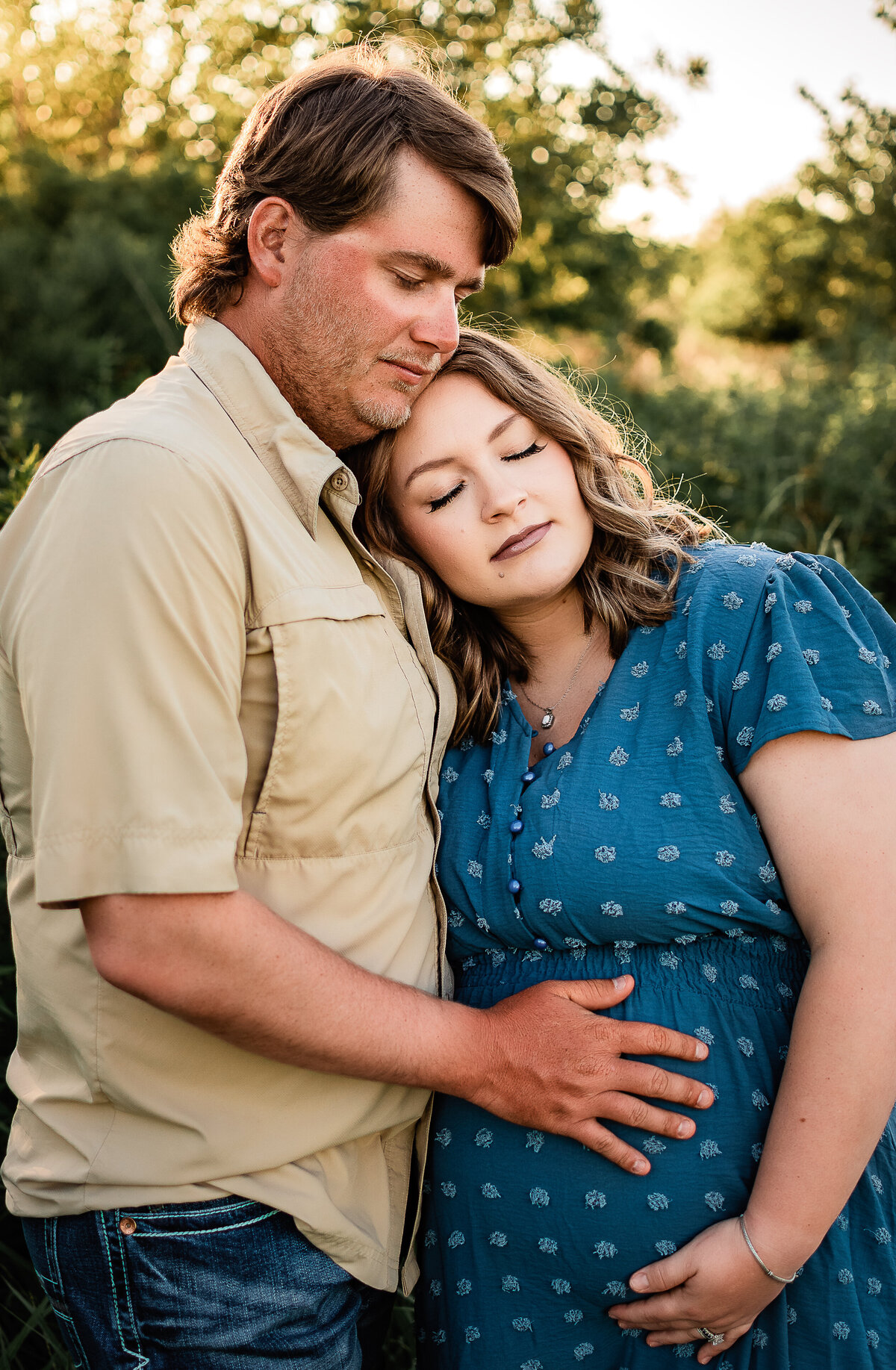 A mom to be in a blue dress rests her head against her husband's chest as they both cradle her bump.