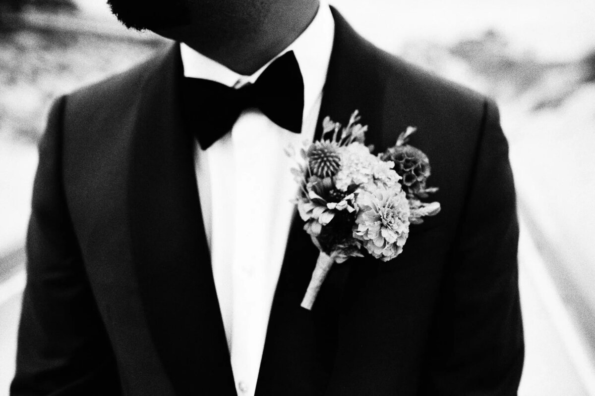 Black-and-white photo by a NJ wedding photographer: a man in a tuxedo with a bow tie and floral boutonniere pinned to his lapel, his face partially out of frame.